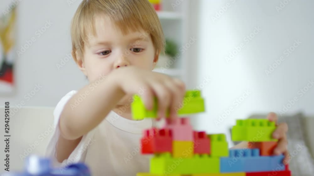 Rack focused shot of cute little boy connecting colorful plastic bricks while playing with construction toy at table Rack focused shot of cute little boy connecting colorful plastic bricks while playing with construction toy at table