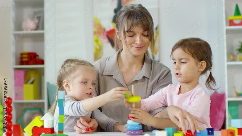 Female teacher smiling and speaking with two cute little girls while they making toy pyramid together at table in kindergarten Female teacher smiling and speaking with two cute little girls while they making toy pyramid together at table in kindergarten