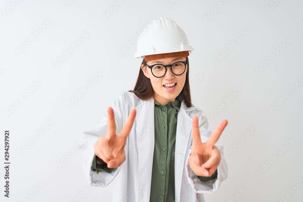 Young chinese engineer woman wearing coat helmet glasses over isolated white background smiling looking to the camera showing fingers doing victory sign. Number two. Young chinese engineer woman wearing coat helmet glasses over isolated white background smiling looking to the camera showing fingers doing victory sign. Number two.