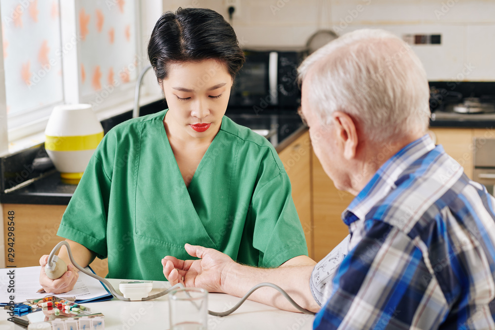 Serious young Asian medical nurse using tonometer to measure blood pressure of senior patient when visiting him at home Serious young Asian medical nurse using tonometer to measure blood pressure of senior patient when visiting him at home