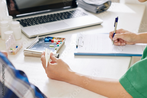 Close-up image of medical nurse checking thermometer and writing down temperature in medical card of senior patient