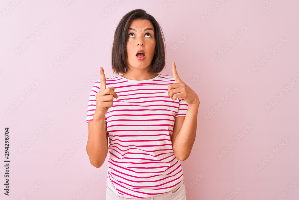 Young beautiful woman wearing striped t-shirt standing over isolated pink background amazed and surprised looking up and pointing with fingers and raised arms. Young beautiful woman wearing striped t-shirt standing over isolated pink background amazed and surprised looking up and pointing with fingers and raised arms.