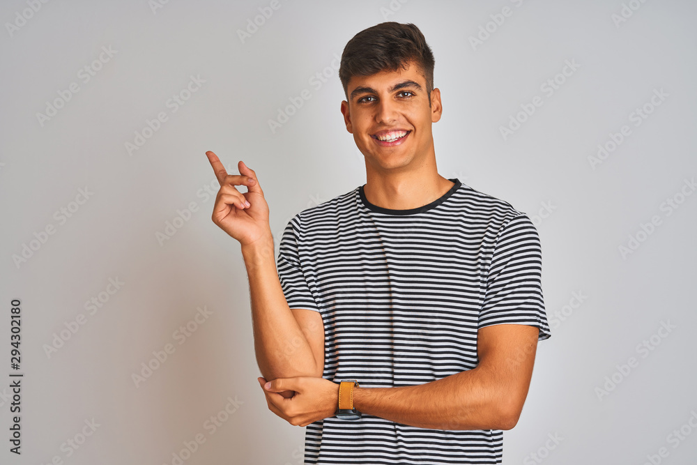 Young indian man wearing navy striped t-shirt standing over isolated white background with a big smile on face, pointing with hand and finger to the side looking at the camera. Young indian man wearing navy striped t-shirt standing over isolated white background with a big smile on face, pointing with hand and finger to the side looking at the camera.