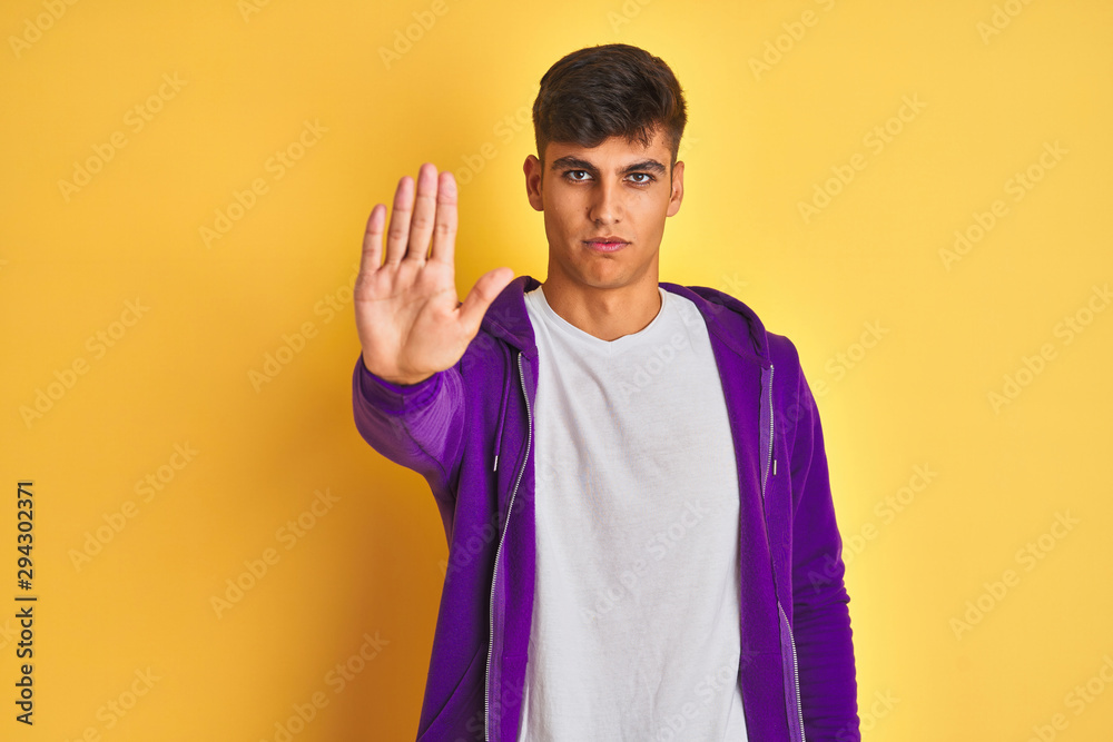 Young indian man wearing purple sweatshirt standing over isolated yellow background doing stop sing with palm of the hand. Warning expression with negative and serious gesture on the face. Young indian man wearing purple sweatshirt standing over isolated yellow background doing stop sing with palm of the hand. Warning expression with negative and serious gesture on the face.
