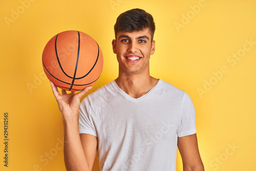 Young indian sportsman holding basketball ball standing over isolated yellow background with a happy face standing and smiling with a confident smile showing teeth
