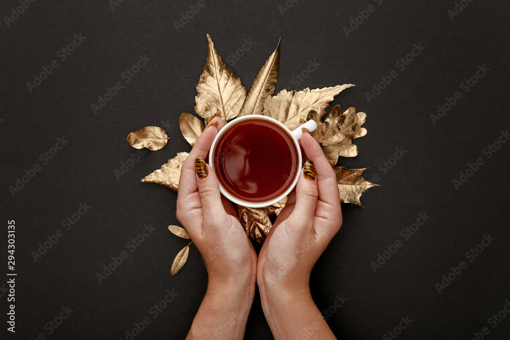 Fototapeta premium partial view of woman holding tea in mug near golden foliage on black background