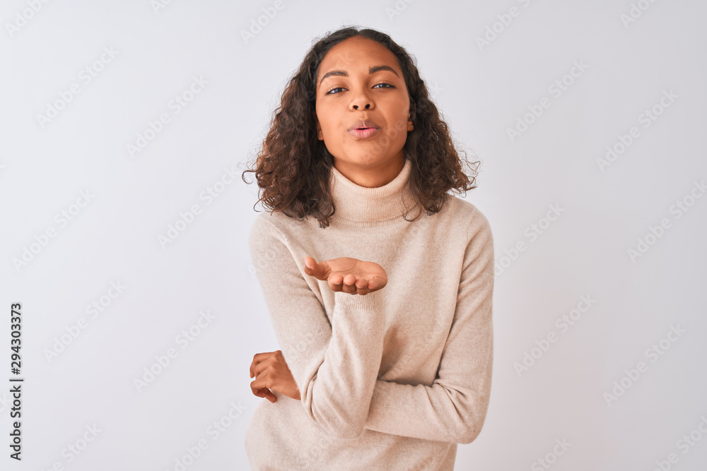 Young brazilian woman wearing turtleneck sweater standing over isolated white background looking at the camera blowing a kiss with hand on air being lovely and sexy. Love expression. Young brazilian woman wearing turtleneck sweater standing over isolated white background looking at the camera blowing a kiss with hand on air being lovely and sexy. Love expression.