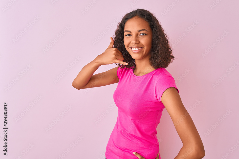 Young brazilian woman wearing t-shirt standing over isolated pink background smiling doing phone gesture with hand and fingers like talking on the telephone. Communicating concepts.