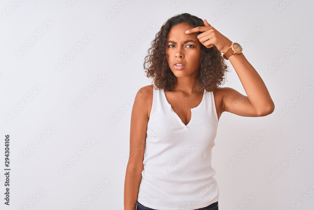 Young brazilian woman wearing casual t-shirt standing over isolated white background pointing unhappy to pimple on forehead, ugly infection of blackhead. Acne and skin problem