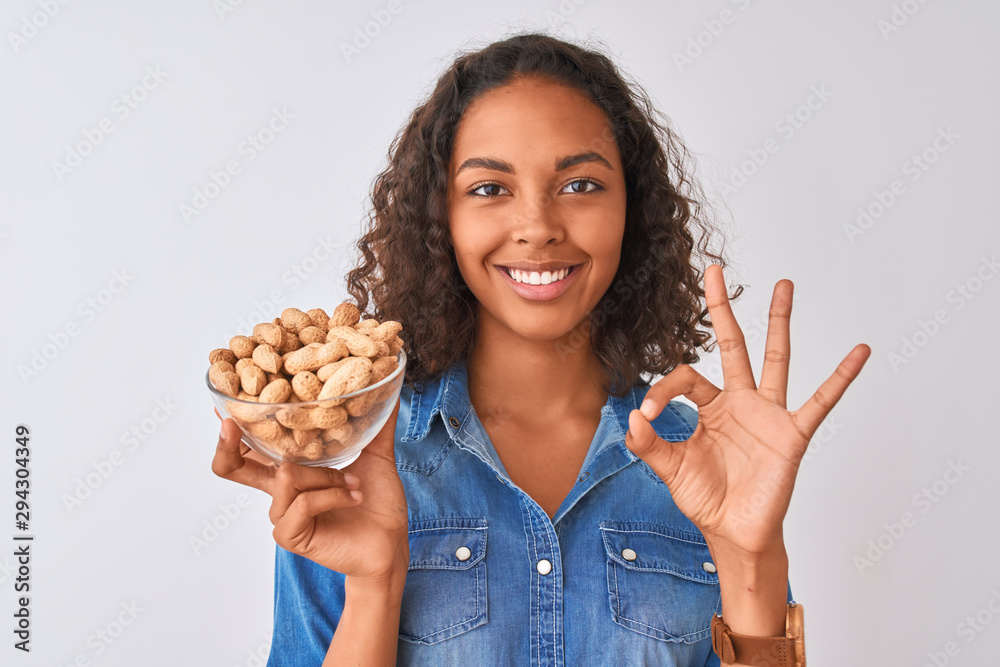 Young brazilian woman holding bowl with peanuts standing over isolated white background doing ok sign with fingers, excellent symbol Young brazilian woman holding bowl with peanuts standing over isolated white background doing ok sign with fingers, excellent symbol