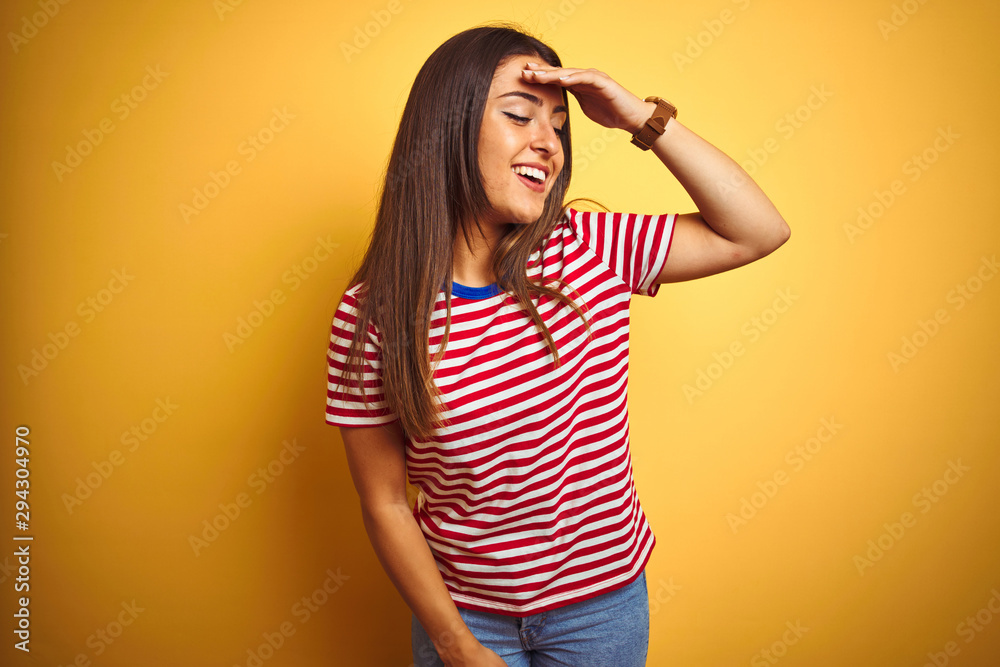 Young beautiful woman wearing striped t-shirt standing over isolated yellow background very happy and smiling looking far away with hand over head. Searching concept. Young beautiful woman wearing striped t-shirt standing over isolated yellow background very happy and smiling looking far away with hand over head. Searching concept.