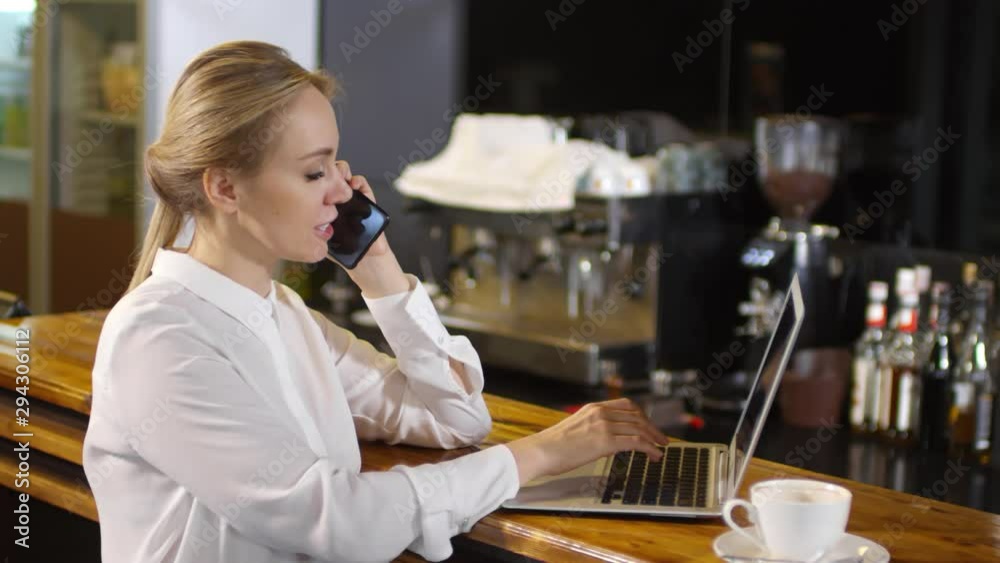 Cheerful caucasian businesswoman in white formal shirt typing on laptop and speaking on mobile phone at the bar counter in cafe Cheerful caucasian businesswoman in white formal shirt typing on laptop and speaking on mobile phone at the bar counter in cafe