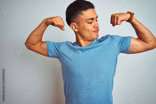 Young brazilian man wearing blue t-shirt standing over isolated white background showing arms muscles smiling proud. Fitness concept.