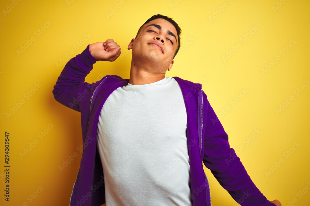 Young brazilian man wearing purple sweatshirt standing over isolated yellow background stretching back, tired and relaxed, sleepy and yawning for early morning