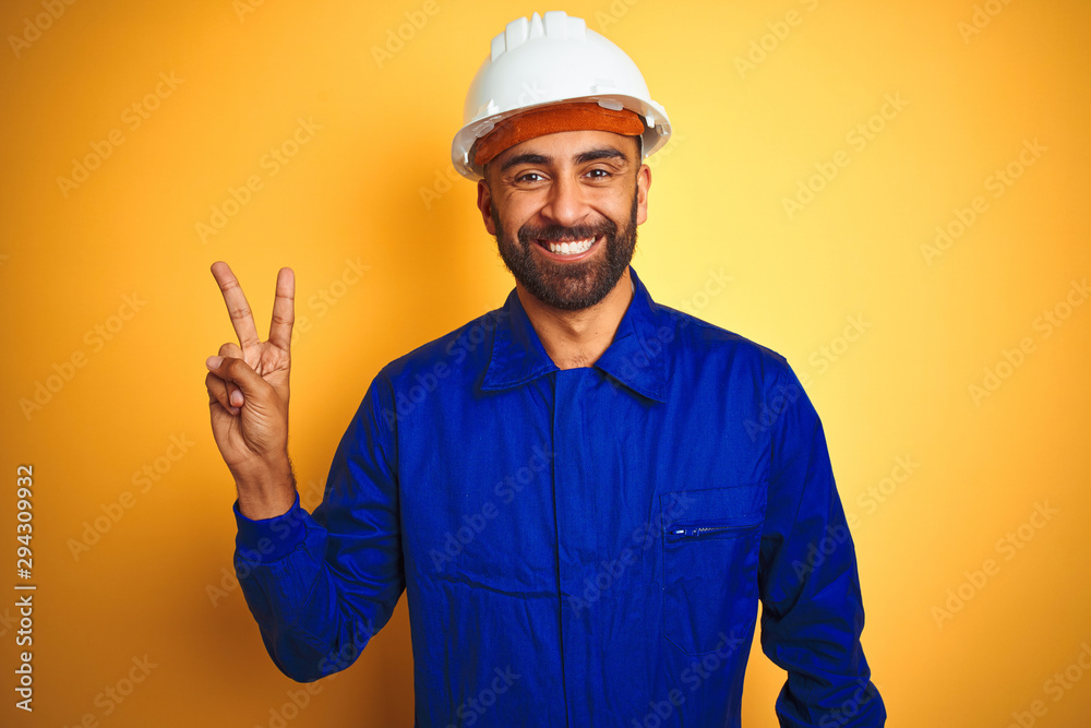 Handsome indian worker man wearing uniform and helmet over isolated yellow background smiling with happy face winking at the camera doing victory sign. Number two.