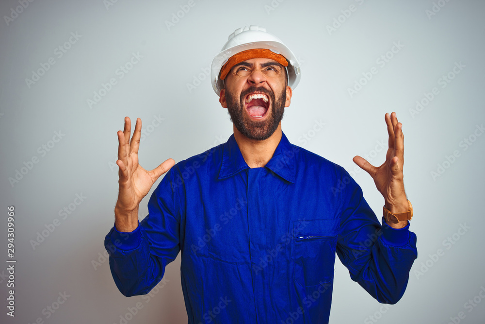 Handsome indian worker man wearing uniform and helmet over isolated white background crazy and mad shouting and yelling with aggressive expression and arms raised. Frustration concept.