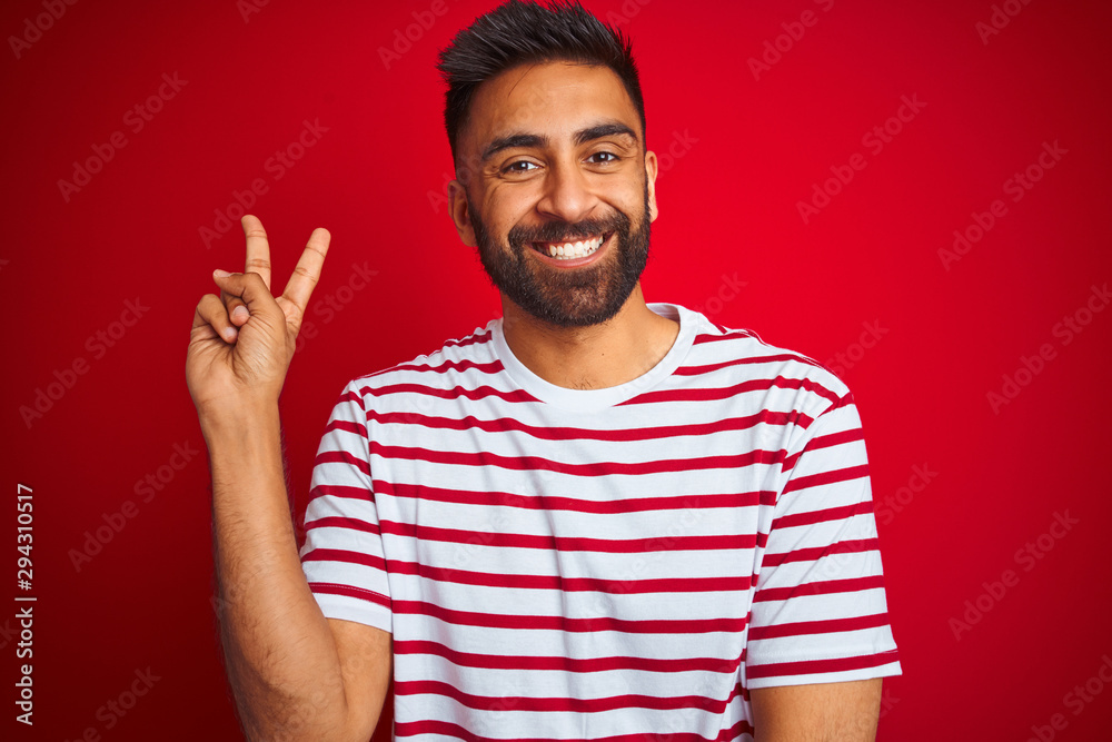 Young indian man wearing striped t-shirt standing over isolated red background smiling with happy face winking at the camera doing victory sign. Number two. Young indian man wearing striped t-shirt standing over isolated red background smiling with happy face winking at the camera doing victory sign. Number two.