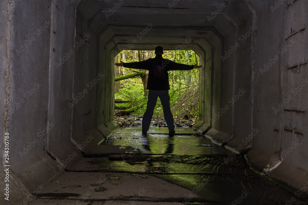Young man standing on end of concrete stream tunnel in black jacket with backpack and open hand Young man standing on end of concrete stream tunnel in black jacket with backpack and open hand