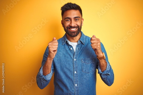 Young indian man wearing denim shirt standing over isolated yellow background excited for success with arms raised and eyes closed celebrating victory smiling. Winner concept.
