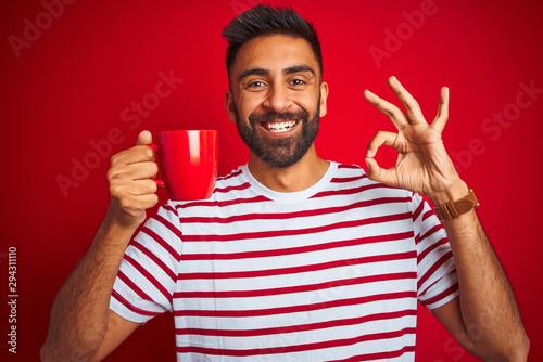 Young indian man wearing striped t-shirt drinking cup of coffee over isolated red background doing ok sign with fingers, excellent symbol © Krakenimages.com