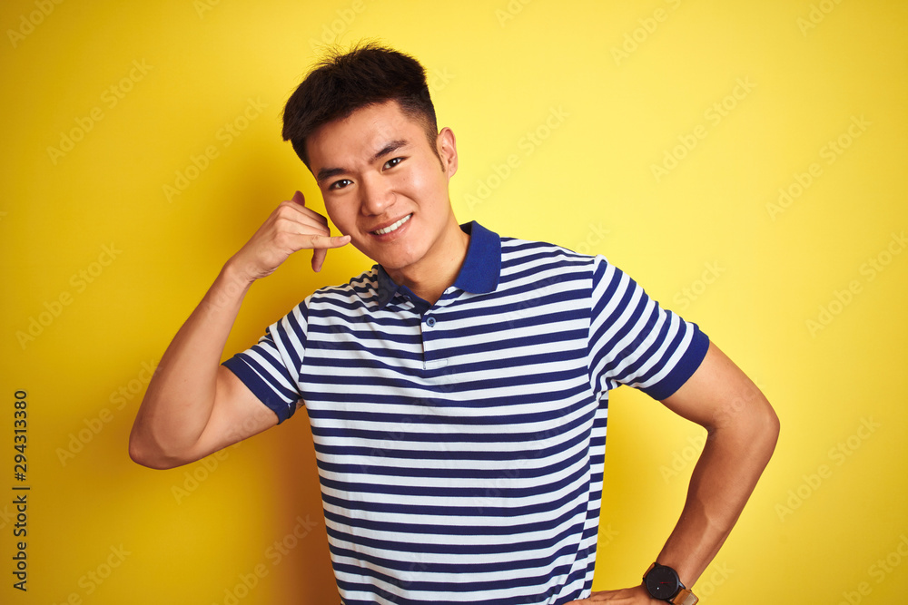 Young asian chinese man wearing striped polo standing over isolated yellow background smiling doing phone gesture with hand and fingers like talking on the telephone. Communicating concepts. Young asian chinese man wearing striped polo standing over isolated yellow background smiling doing phone gesture with hand and fingers like talking on the telephone. Communicating concepts.
