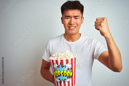 Young asian chinese man holding pack of popcorn standing over isolated white background annoyed and frustrated shouting with anger, crazy and yelling with raised hand, anger concept photo