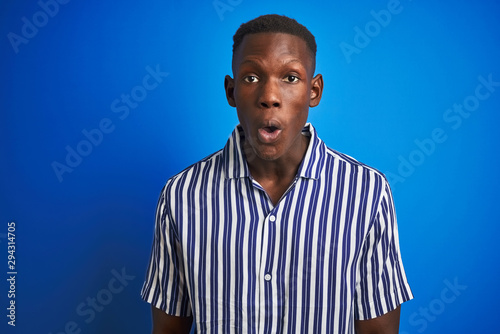 African american man wearing striped casual shirt standing over isolated blue background afraid and shocked with surprise expression, fear and excited face.