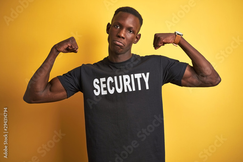 African american safeguard man wearing security uniform over isolated yellow background showing arms muscles smiling proud. Fitness concept.