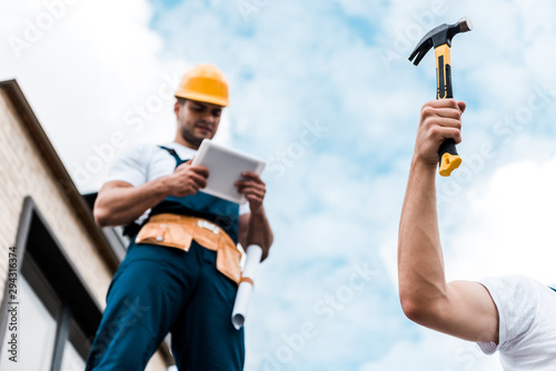 selective focus of man holding hammer near coworker in helmet using digital tablet