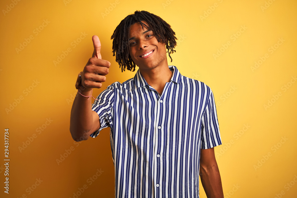 Afro man with dreadlocks wearing casual striped t-shirt over isolated yellow background doing happy thumbs up gesture with hand. Approving expression looking at the camera showing success. Afro man with dreadlocks wearing casual striped t-shirt over isolated yellow background doing happy thumbs up gesture with hand. Approving expression looking at the camera showing success.