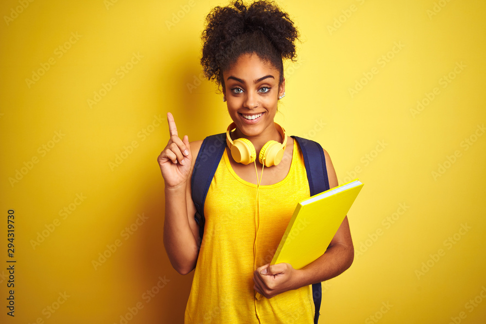 Afro woman using backpack and headphones holding notebook over isolated yellow background surprised with an idea or question pointing finger with happy face, number one Afro woman using backpack and headphones holding notebook over isolated yellow background surprised with an idea or question pointing finger with happy face, number one