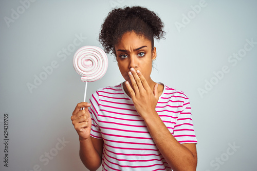 African american teenager woman eating colorful candy over isolated white background cover mouth with hand shocked with shame for mistake, expression of fear, scared in silence, secret concept