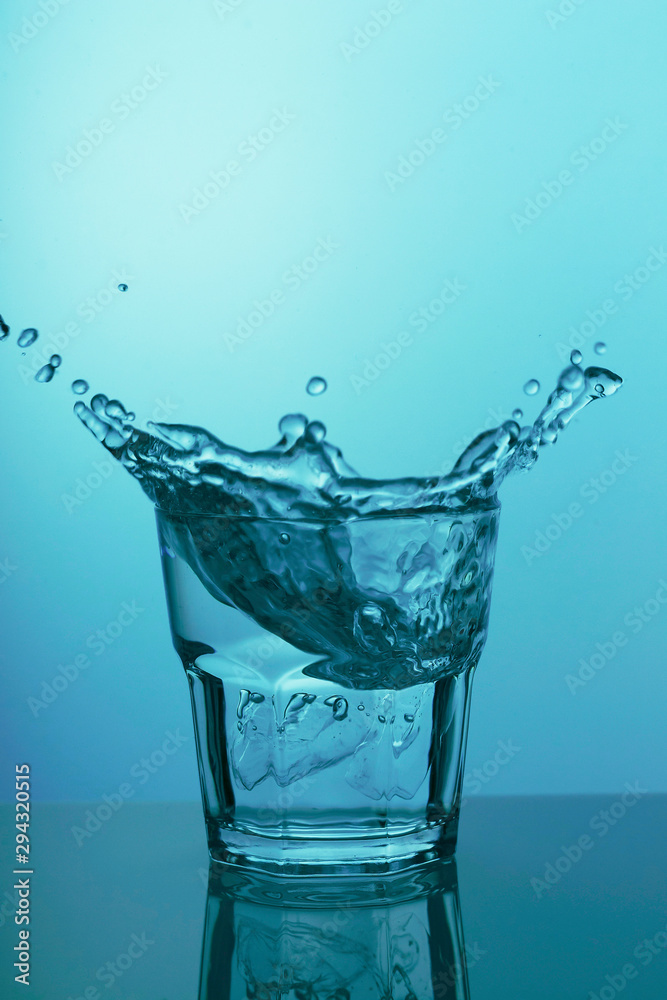 Glass with clear water and splashes on blue background Glass with clear water and splashes on blue background