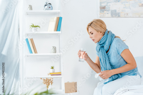 selective focus of sick woman looking at glass of water while holding napkin in bedroom