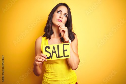 Young beautiful woman holding sale poster over yellow isolated background serious face thinking about question, very confused idea
