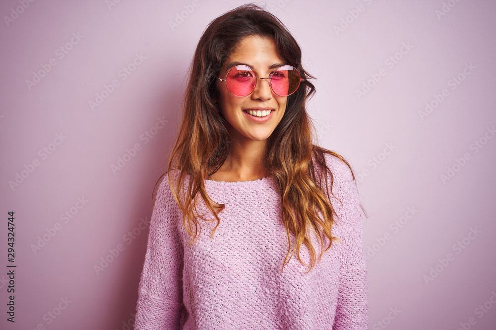Young beautiful woman wearing fashion sunglasses standing over pink isolated background looking away to side with smile on face, natural expression. Laughing confident. Young beautiful woman wearing fashion sunglasses standing over pink isolated background looking away to side with smile on face, natural expression. Laughing confident.
