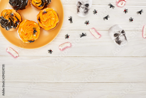 top view of cupcakes on orange plate, gummy teeth, skulls and spiders on white wooden table, Halloween treat