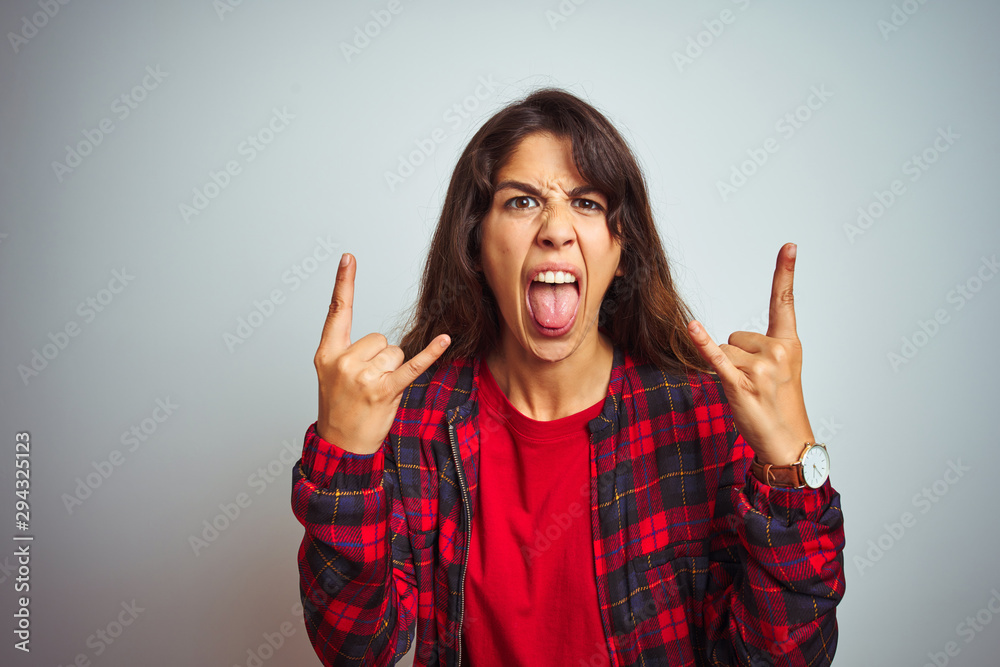 Young beautiful woman wearing red t-shirt and jacket standing over white isolated background shouting with crazy expression doing rock symbol with hands up. Music star. Heavy concept. Young beautiful woman wearing red t-shirt and jacket standing over white isolated background shouting with crazy expression doing rock symbol with hands up. Music star. Heavy concept.