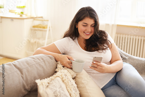 Technology, communication and leisure concept. Pretty girl with chubby cheeks relaxing at home with electronic gadget. Overweight young brunette woman texting sms on smart phone and having coffee photo
