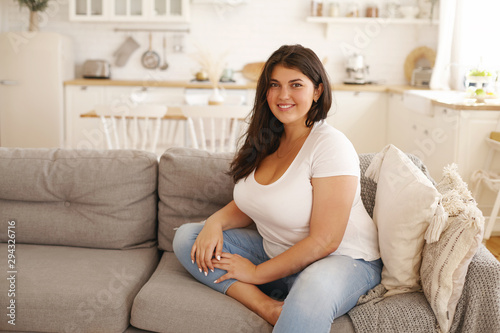 Relaxation, leisure, rest, domesticity and coziness. Beautiful charming young female with chubby cheeks and curvy body sitting on sofa barefooted, keeping one foot on floor, smiling at camera photo