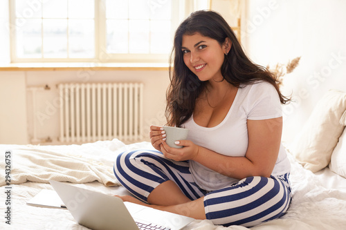 Foto Indoor image of cute curvaceous young brunette woman having cup of coffee in bedroom, sitting on bed with window in background