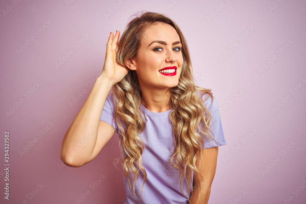 Young beautiful woman wearing purple t-shirt standing over pink isolated background smiling with hand over ear listening an hearing to rumor or gossip. Deafness concept. Young beautiful woman wearing purple t-shirt standing over pink isolated background smiling with hand over ear listening an hearing to rumor or gossip. Deafness concept.