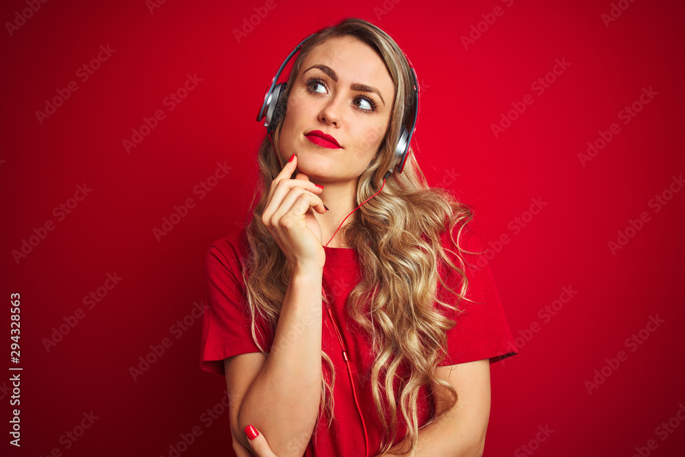 Young beautiful woman wearing headphones over red isolated background with hand on chin thinking about question, pensive expression. Smiling with thoughtful face. Doubt concept. Young beautiful woman wearing headphones over red isolated background with hand on chin thinking about question, pensive expression. Smiling with thoughtful face. Doubt concept.