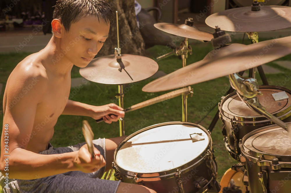Obraz premium Asian American teenager playing drums. Summer portrait of handsome young boy practicing on drum kit at home garden rehearsing rock song enjoying his hobby
