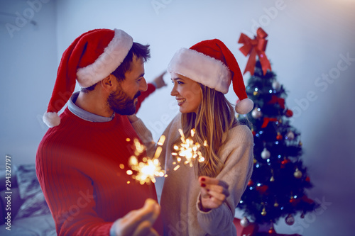 Happy caucasian attractive couple congratulating the new year to each other. Couple having santa hats on heads and holding sprinklers while standing in living room. In background is christmas tree.