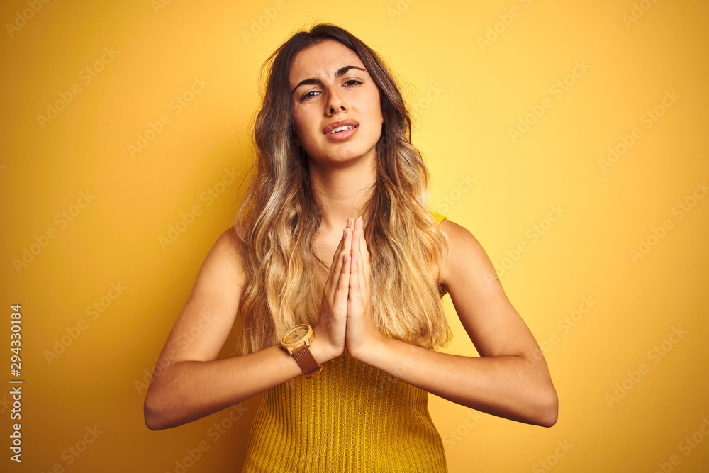 Young beautiful woman wearing t-shirt over yellow isolated background begging and praying with hands together with hope expression on face very emotional and worried. Asking for forgiveness