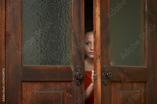 Fotografía A little scared girl is looking through the door slit