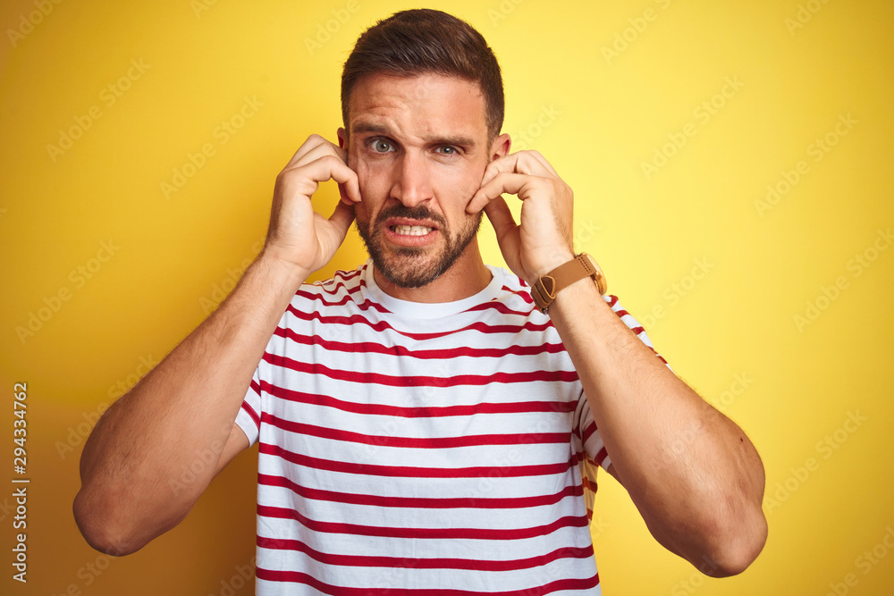 Young handsome man wearing casual red striped t-shirt over yellow isolated background covering ears with fingers with annoyed expression for the noise of loud music. Deaf concept. Young handsome man wearing casual red striped t-shirt over yellow isolated background covering ears with fingers with annoyed expression for the noise of loud music. Deaf concept.