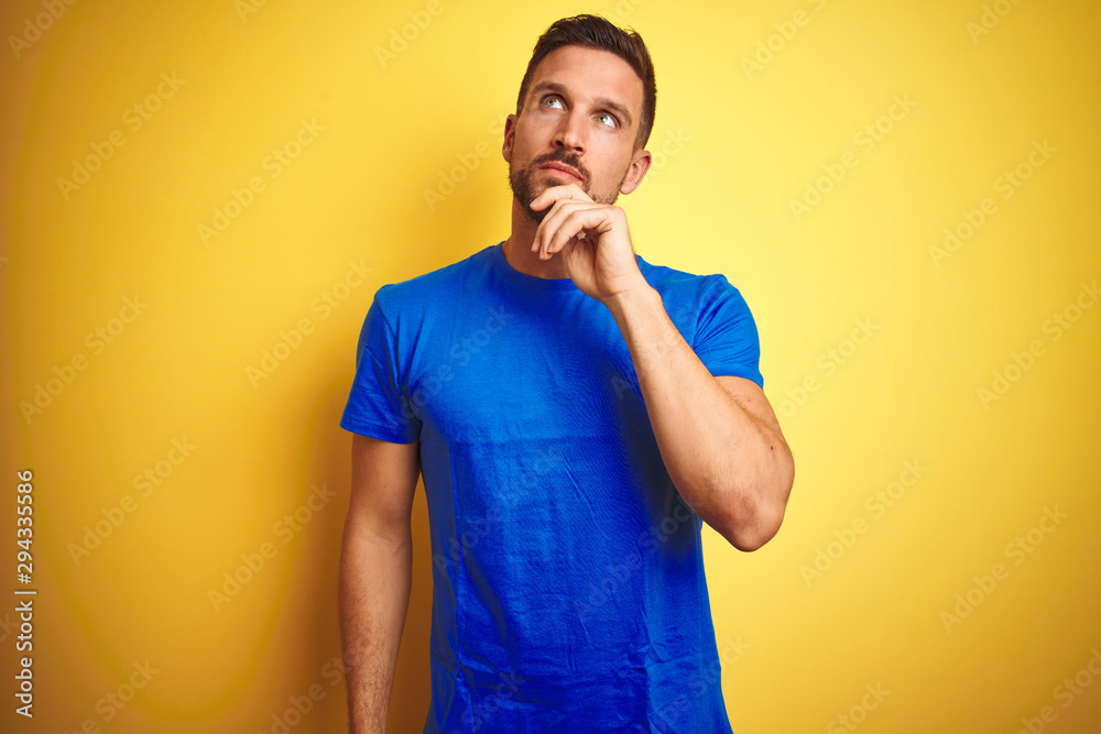 Young handsome man wearing casual blue t-shirt over yellow isolated background with hand on chin thinking about question, pensive expression. Smiling with thoughtful face. Doubt concept. Young handsome man wearing casual blue t-shirt over yellow isolated background with hand on chin thinking about question, pensive expression. Smiling with thoughtful face. Doubt concept.