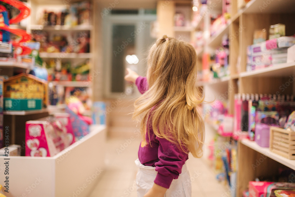 Back view of adorable little girl shopping for toys. Cute female in toy store. Happy young girl selecting toy